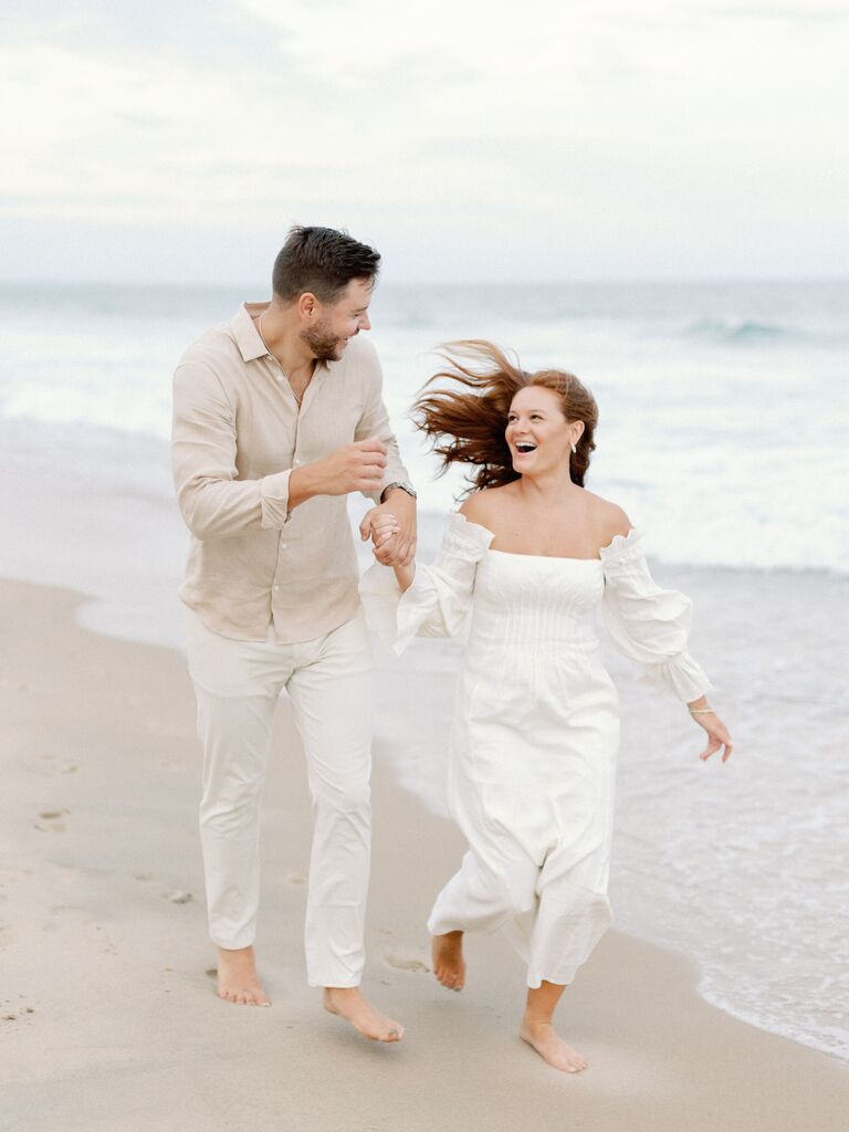 Couple holds hands while running down a beach. 