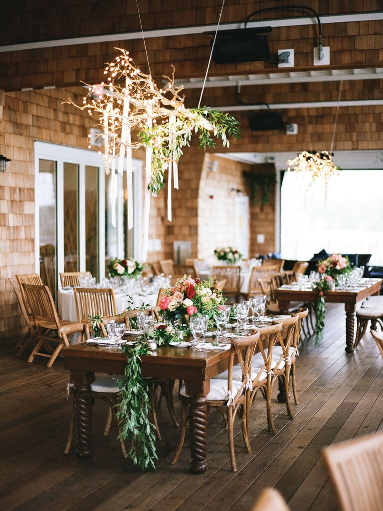 rustic indoor wedding reception decor with string lights and greenery on a tree branch hanging above long wooden table