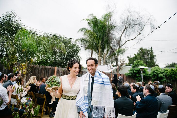 Emily and Chris walked up the aisle after their Jewish wedding ceremony, with the tallith draped around Chris's shoulders. Emily wore a simple necklace and dangling earrings on the wedding day, which accented her ivory dress with a green-fabric flower belt.