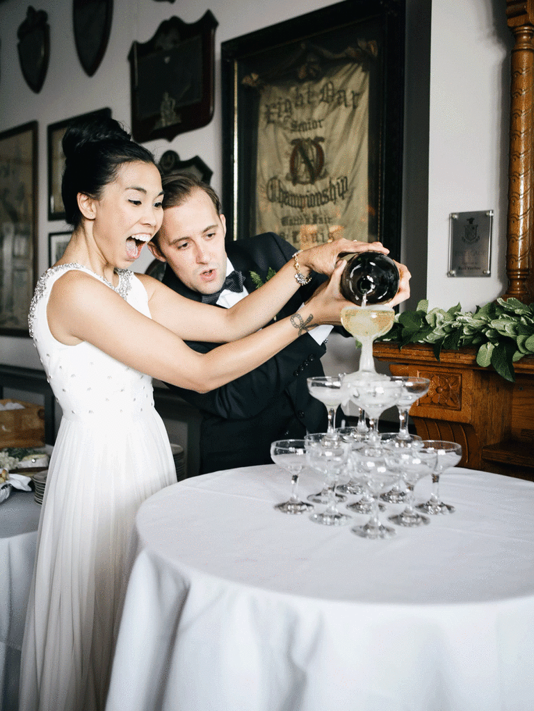 Bride and groom pop a bottle of champagne and poured it over a wedding champagne tower. 