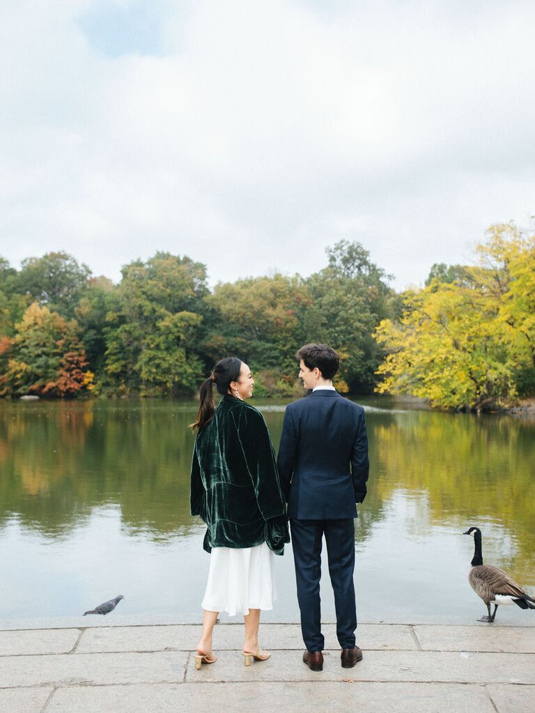 A couple stand at a pond together with some Canada geese