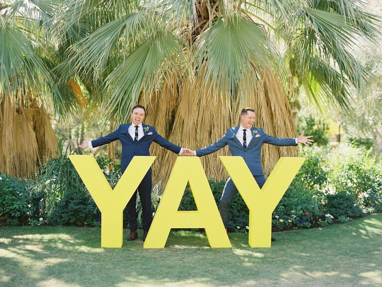 Couple poses with a large sign that reads YAY. 