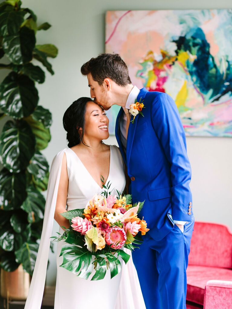 Groom kissing bride holding colorful bouquet of tropical flowers