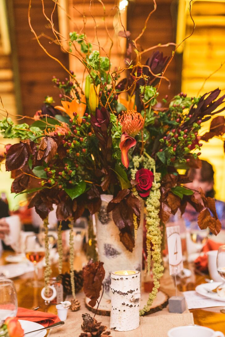 Wooden tables at the reception were paired with one of two centerpieces; the tallest versions took natural to a new level. Birch wood formed the vase while a display of greenery, red hypericum berries, magnolia leaves, mango calla lilies, red roses, curly willow and orange tulips spilled over the sides. Matching birch-wood-accented candles and pinecones also circled the arrangements.