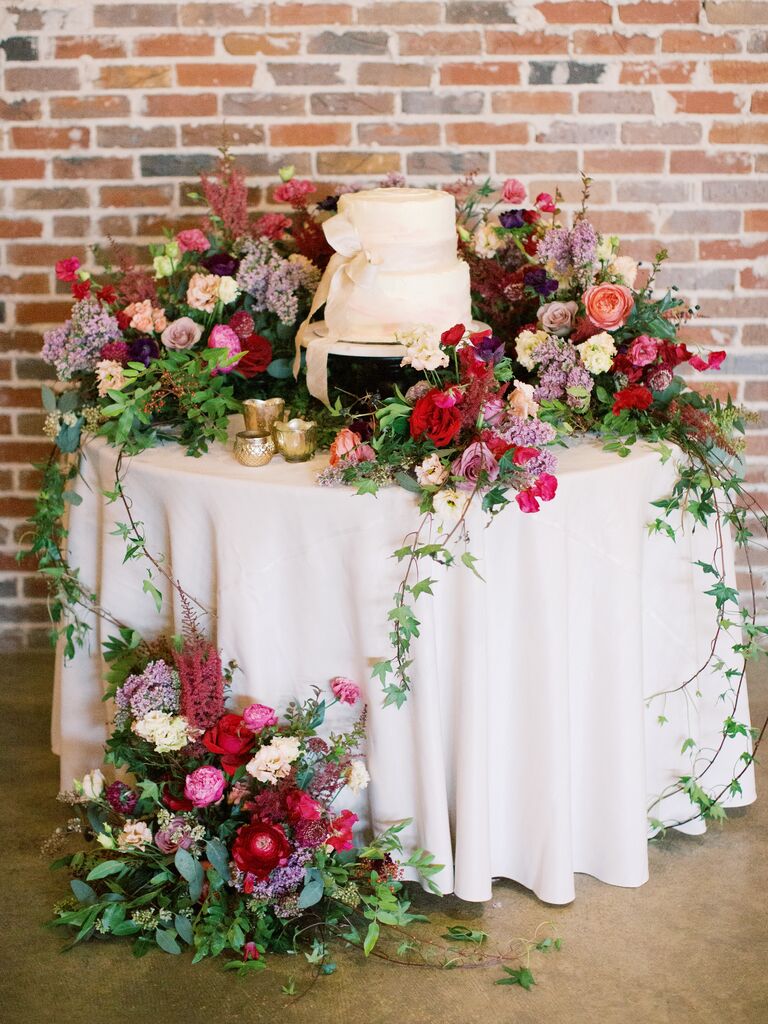 wedding cake displayed on a round table overflowing with colorful pink and purple flowers and vines cascading to the floor