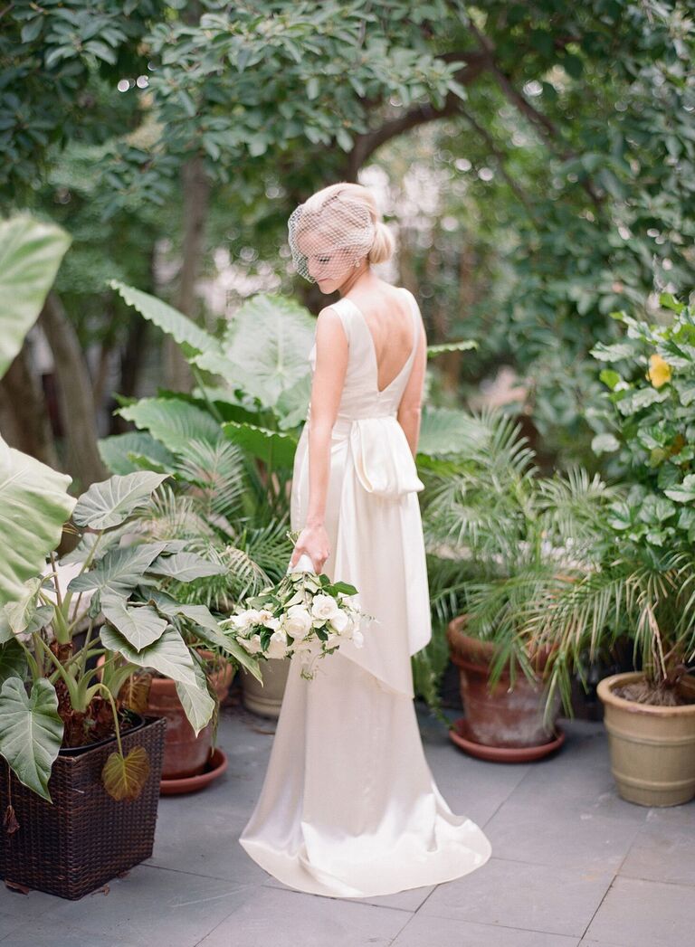 bride wearing satin sheath gown and birdcage veil stands among potted plants at garden venue