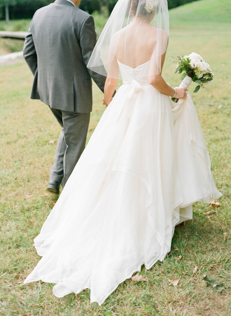 bride wearing tulle elbow length wedding veil holds hands with groom and walks across grass