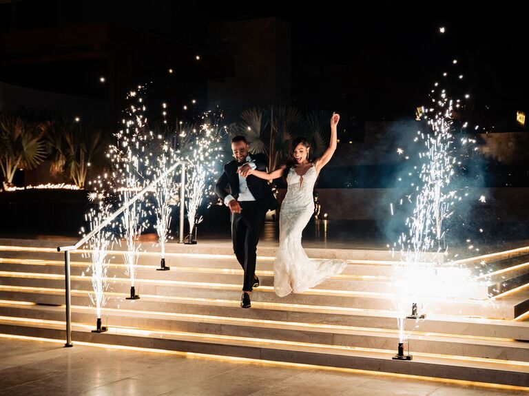 Bride and groom walking down stairs with sparklers machines next to them.