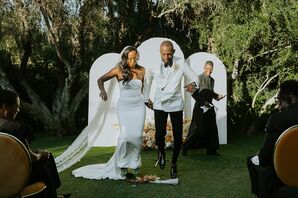 Bride in Fitted, Sleek Gown and Long Veil and Groom in White Tux Jacket Jump the Broom, Altar Backdrop With Flowers