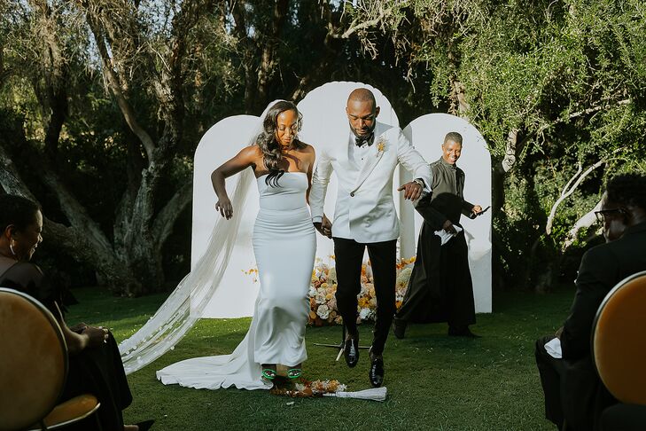 Bride in Fitted, Sleek Gown and Long Veil and Groom in White Tux Jacket Jump the Broom, Altar Backdrop With Flowers