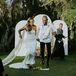 Bride in Fitted, Sleek Gown and Long Veil and Groom in White Tux Jacket Jump the Broom, Altar Backdrop With Flowers