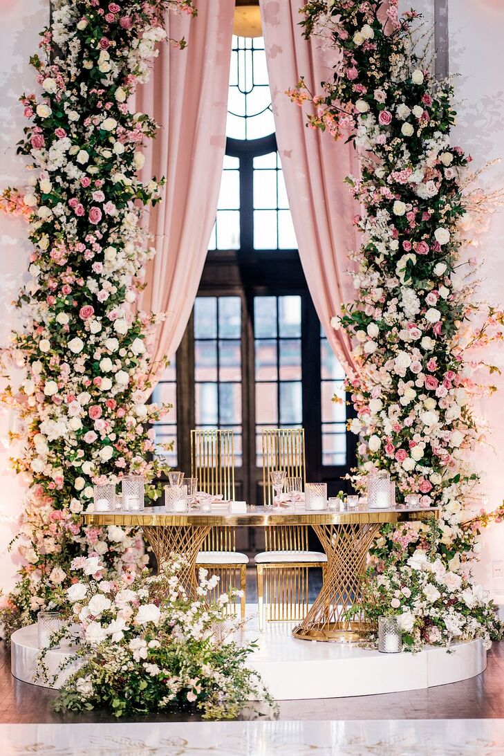 A gold sweetheart table at a couple's modern-fairytale-themed wedding reception featuring climbing pink flowers and greenery, floral arrangements spilling onto the floor, pink drapery and tons of candles.
