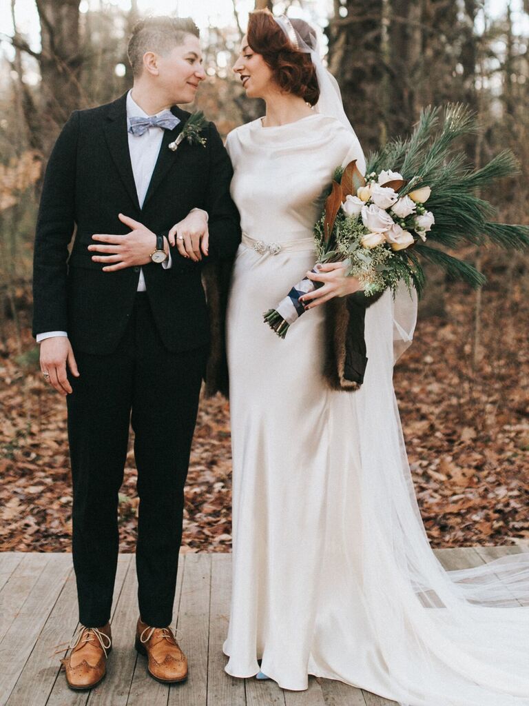 couple standing outside in front of forest with bride holding pageant style wedding bouquet of winter evergreens and roses