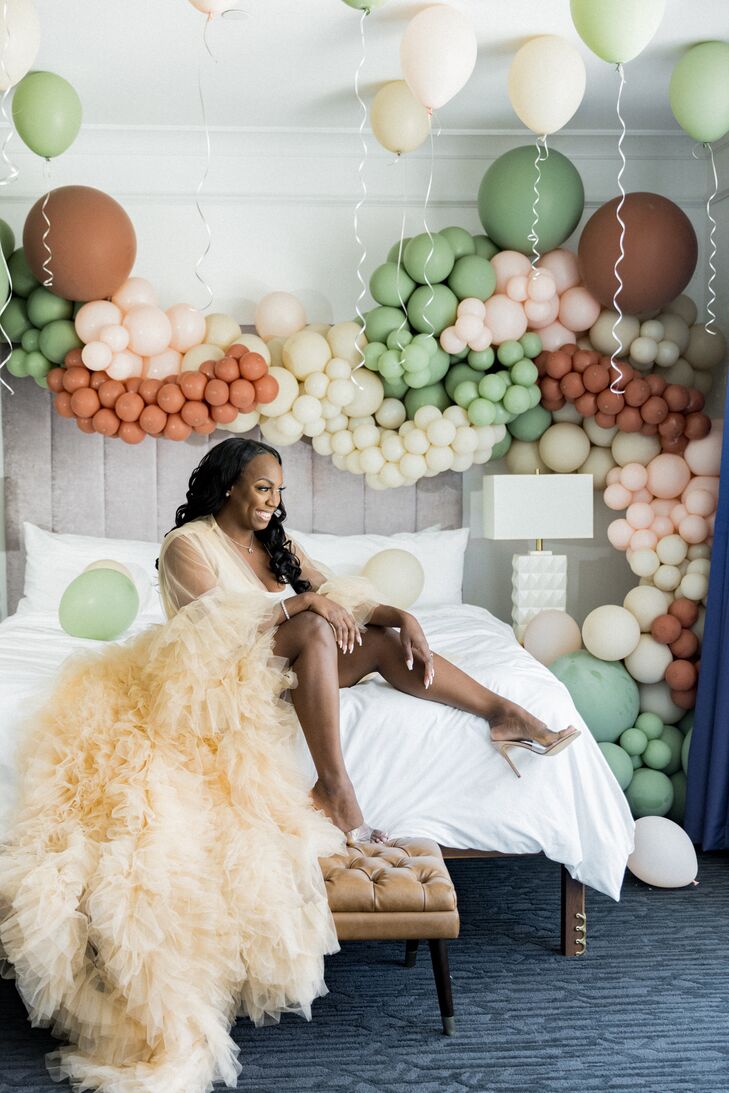 A bride getting ready in a glamorous, champagne-colored robe with tulle ruffles in a hotel room with green and burnt orange balloons and high heels before her bohemian-chic wedding.