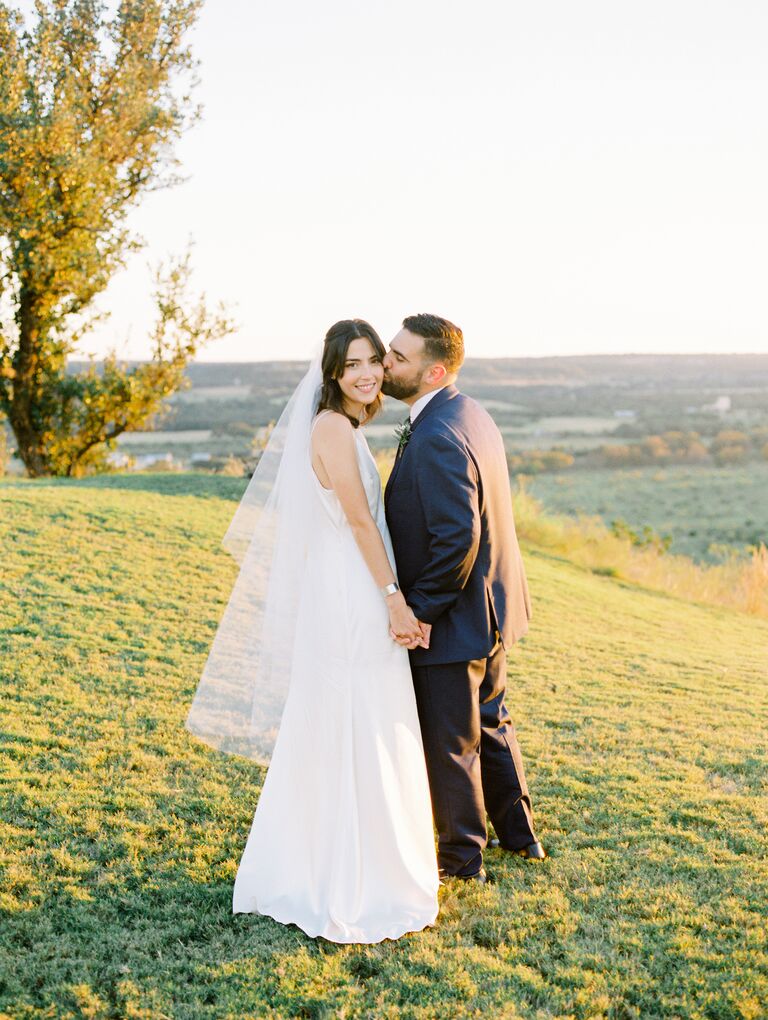 bride wearing modern satin sheath wedding dress with tulle ballet length wedding veil holds grooms hand and looks at the camera as he kisses her on the cheek