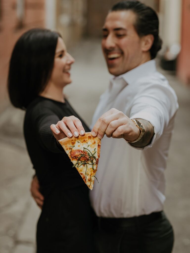 Couple holds out a slice of pizza while embracing. 