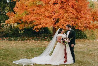 A couple pose in their wedding attire in front of a stunning autumn-toned tree.