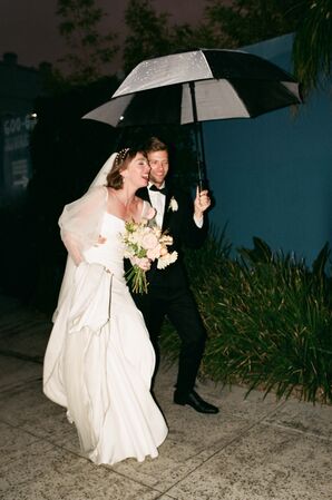 Bride With Trendy Wedding Gown, Light Bouquet and Groom in Classic Tuxedo Together Under Umbrella