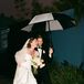 Bride With Trendy Wedding Gown, Light Bouquet and Groom in Classic Tuxedo Together Under Umbrella