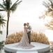 A bride in a luxurious, bohemian wedding gown featuring an off-the-shoulder neckline, lace details and a champagne-colored underlay with a gold crown to represent her Greek heritage kisses her groom in a light gray suit without a jacket on their ceremony altar platform in front of a pampas grass semi-circle overlooking the ocean in Phuket, Thailand.