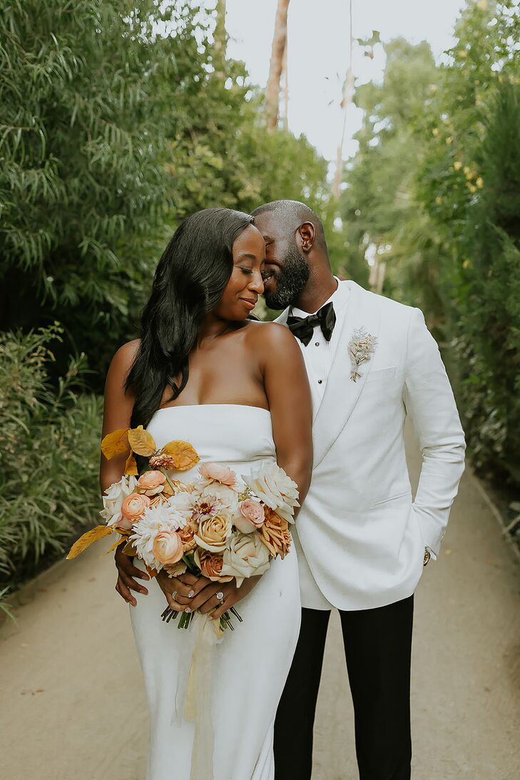 A bride in a sleek, fitted, strapless gown without adornments and a relaxed, down hairstyle holds a bouquet of orange, coral, dusty rose and ivory blossoms while embracing her groom in a white tuxedo jacket and black pants and a simple boutonniere in a garden at their desert venue.