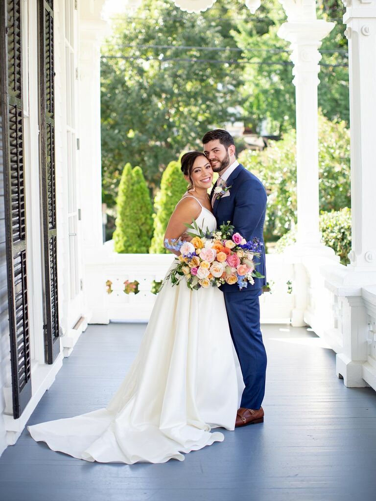 Bride and groom embrace while holding a beautiful bouquet featuring dahlias and other fresh flowers. 