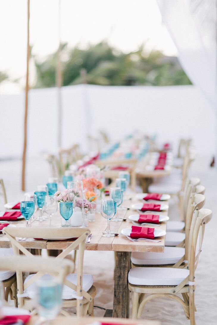 Dining tables and chairs in light wood, set up on the sand, had turquoise glasses, pops of pink and coral flowers, which added to Danielle and Jason’s rustic, casual reception vibe.