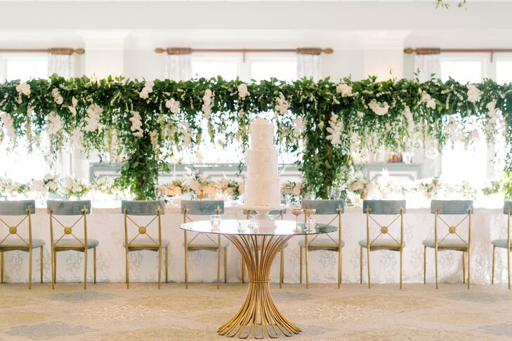 Cake on Gold-and-Glass Table in Front of Greenery-Accented Reception Tables