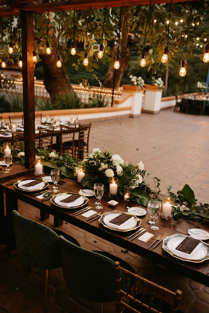 A long, wood head table for the grooms and their wedding party with a suspended installation overhead featuring hanging Edison bulbs and greenery above candles, low centerpieces and gold-rimmed chargers with custom menus at a black-tie, outdoor wedding reception.