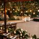 A long, wood head table for the grooms and their wedding party with a suspended installation overhead featuring hanging Edison bulbs and greenery above candles, low centerpieces and gold-rimmed chargers with custom menus at a black-tie, outdoor wedding reception.