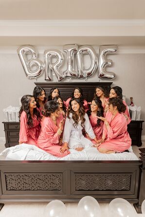 Bride in White, Silk Robe Surrounded by Bridesmaids in Pink Robes on Bed With Balloons in Hotel