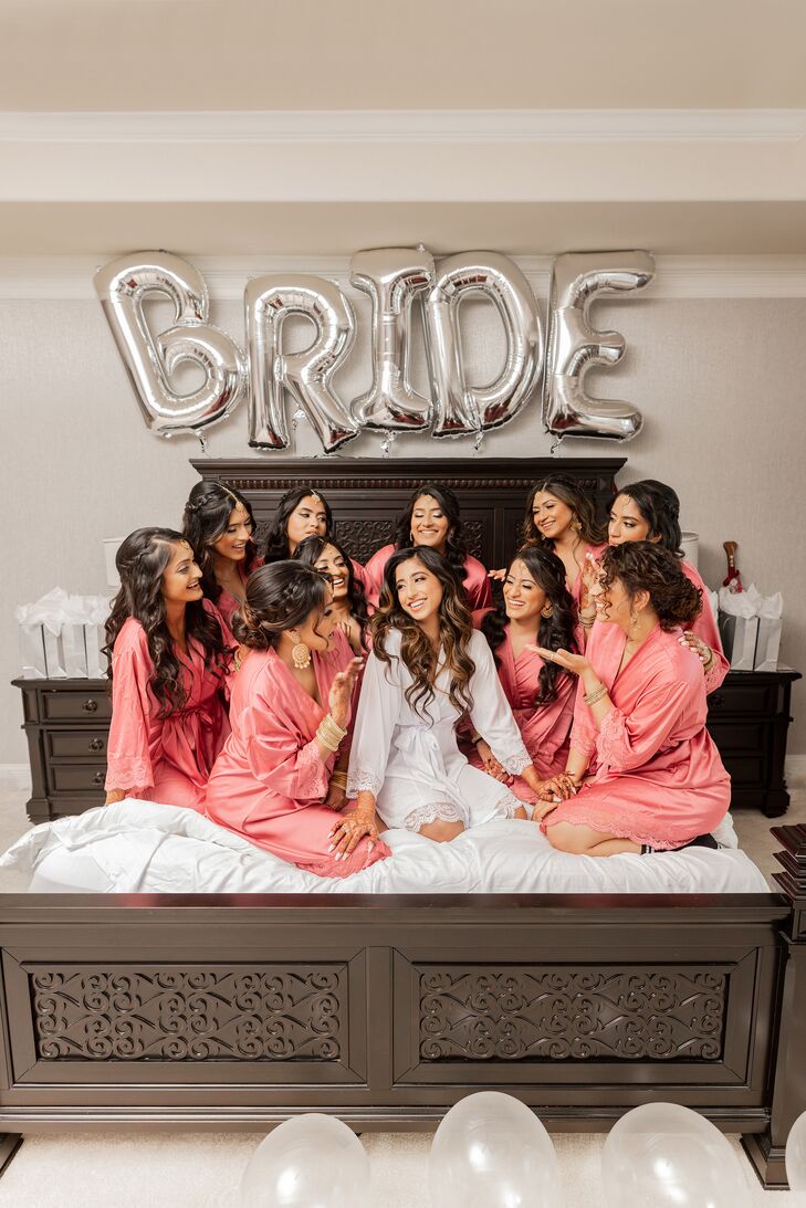 Bride in White, Silk Robe Surrounded by Bridesmaids in Pink Robes on Bed With Balloons in Hotel