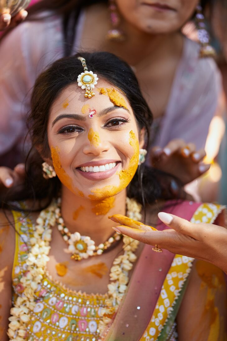 An Indian bride with a bejeweled Bindi headpiece, luxurious earrings and necklaces getting yellow turmeric put on her face at a prewedding event called a Haldi ceremony.