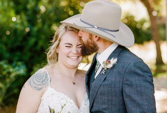 Man wearing a cowboy hat gives a sweet kiss on the cheek to his smiling bride. 