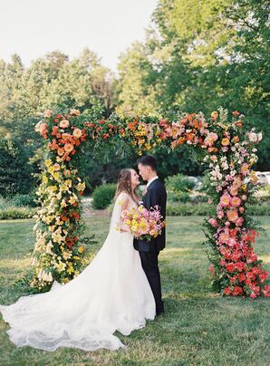 Bride With Long Cape Trail and Groom With Colorful Bouquet Under Lush Arch