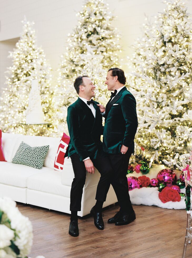 Two grooms in coordinating emerald green velvet suit jackets bow ties with different lapels standing in their Christmas wedding reception lounge space in front of Christmas trees, large ornaments, faux snow and colorful bouquets.