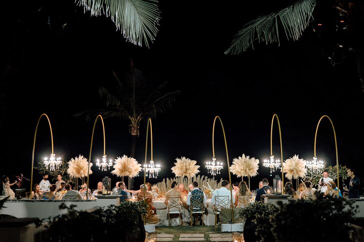 A beautiful nighttime scene of a small wedding reception with one long dining table featuring tall pampas grass centerpieces between crescent floor stands with hanging chandeliers under a starry night sky and palm trees at an outdoor, beachfront wedding in Phuket, Thailand.