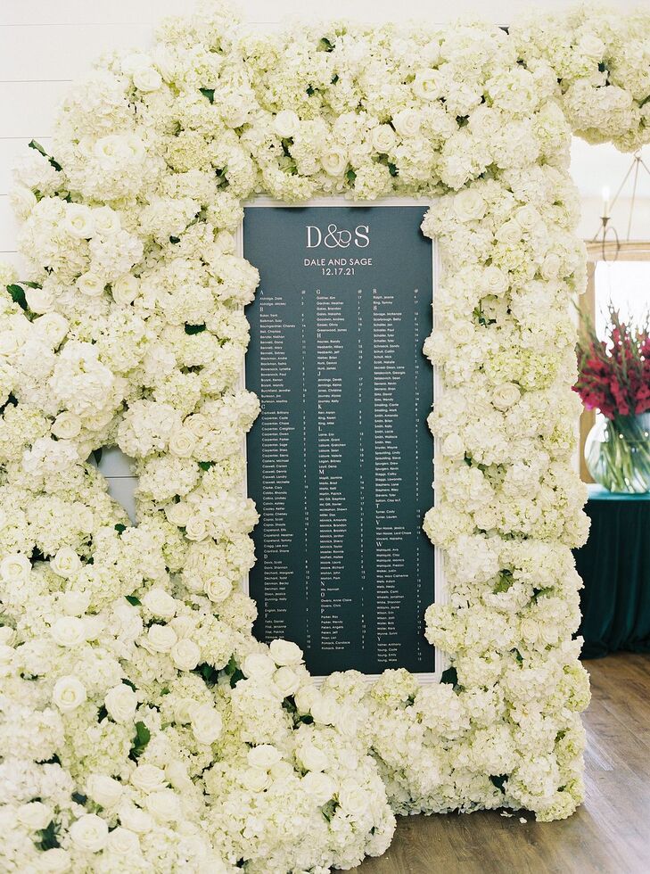 A cold emerald green seating chart surrounded by a lush flower wall of white hydrangeas, garden roses and other crisp winter blossoms at a Christmas wedding reception.