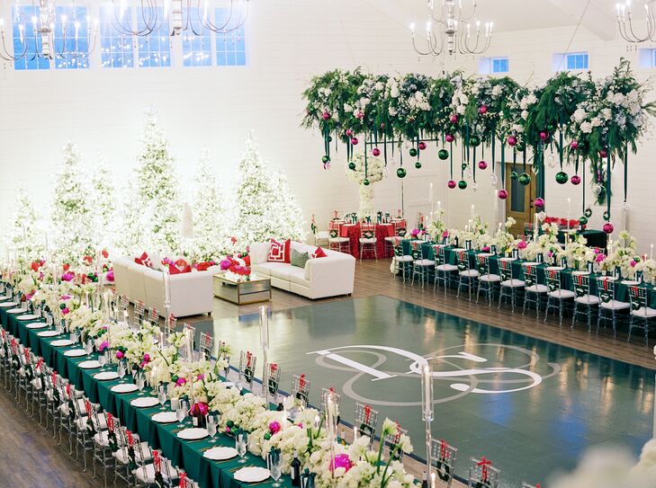 A bold, whimsical Christmas wedding reception with a custom dance floor, white lounge space, Christmas trees with lights, long and round dining tables with emerald green and fuchsia linens and long table runners all sitting under a hanging arrangement of greenery, white flowers and vibrant suspended ornaments.