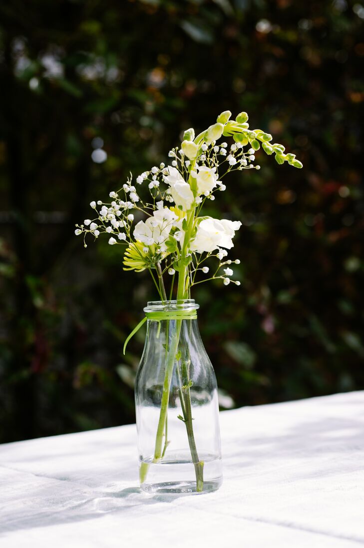 At the backyard reception, dining tables dressed in simple white linens were decorated with glass milk bottle used as vases, filled with green button chrysanthemums and white stock accented with baby's breath. The top of the glass bottle was wrapped with green ribbon.