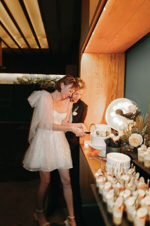 Bride in Short Dress, Shoulder Bows and Groom in Tux Cutting Small Wedding Cake, Dessert Table