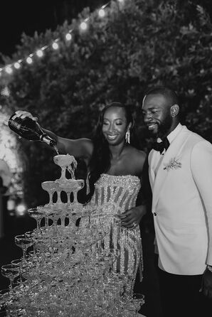 Black-and-White Photo of Bride in Sparkly Mini Dress and Groom in White Tux Jacket Pouring Champagne on Tower