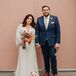 A bride in a long-sleeve, v-neck sequin gown and wavy down hairstyle with a bright, warm-toned bouquet with orange, red, yellow, pink and blue flowers with her groom in a navy suit and bright boutonniere and pocket square standing with their dog in a mini tuxedo shirt.