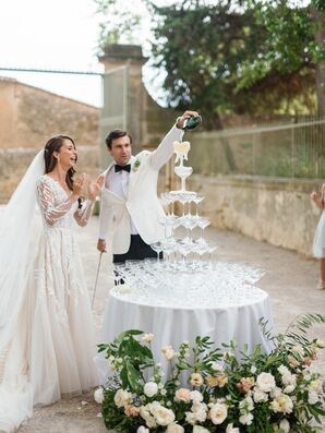 Bride in Elegant Gown, Long Veil Watches as Groom in White Tux Jacket Pours Champagne on Tower of Glasses