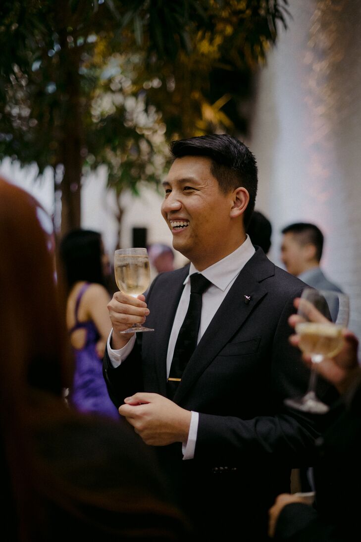 Groom in Classic Black-and-White Suit Smiling, Watching Toasts With Drink in Hand