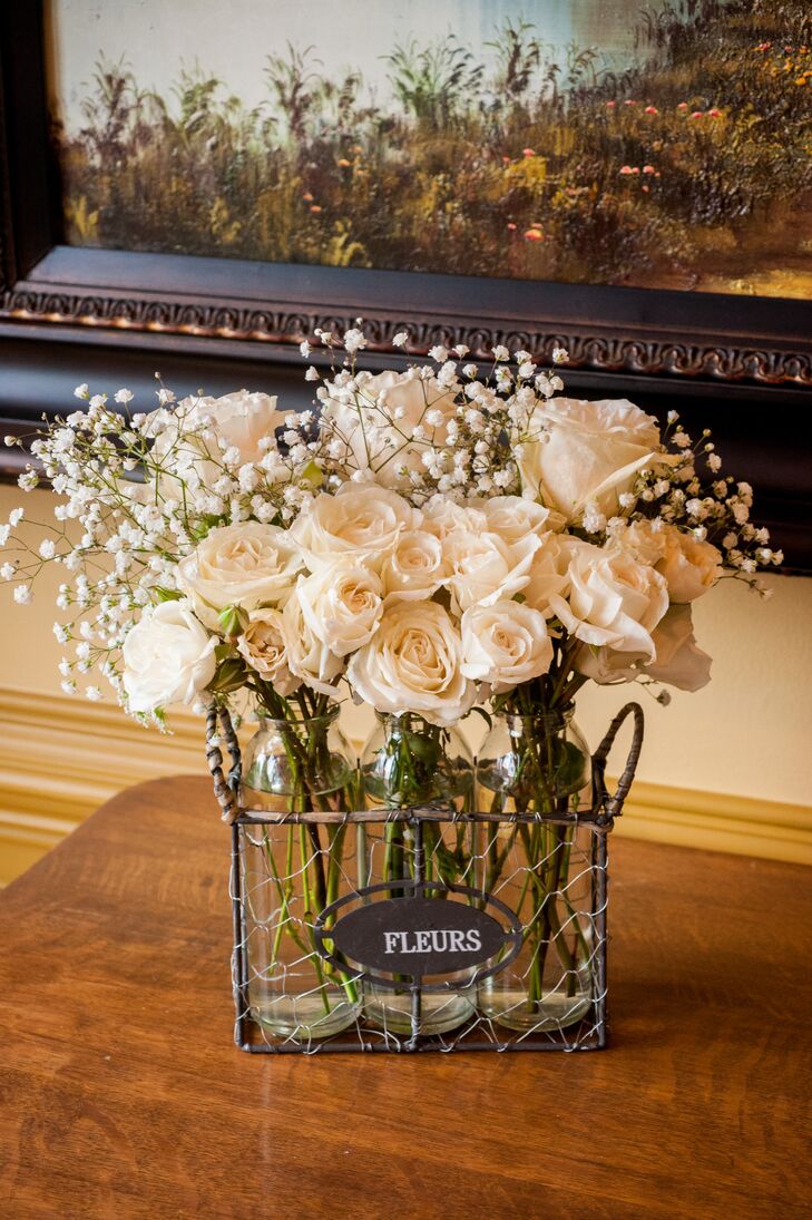 Elegant ivory roses mixed with baby's breath inside an iron container used for decorative means, with a black-and-white sign on the front that read "fleurs."
