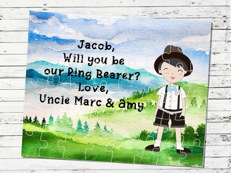A jigsaw puzzle with mountainscape art and a child that reads "Jacob, will you be our ring bearer?" from Etsy