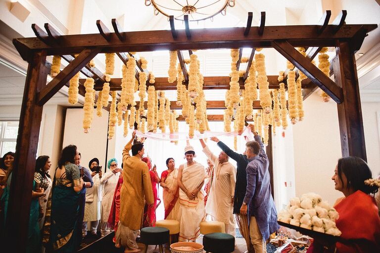 Wedding ceremony taking place beneath wood mandap with orange flower accents