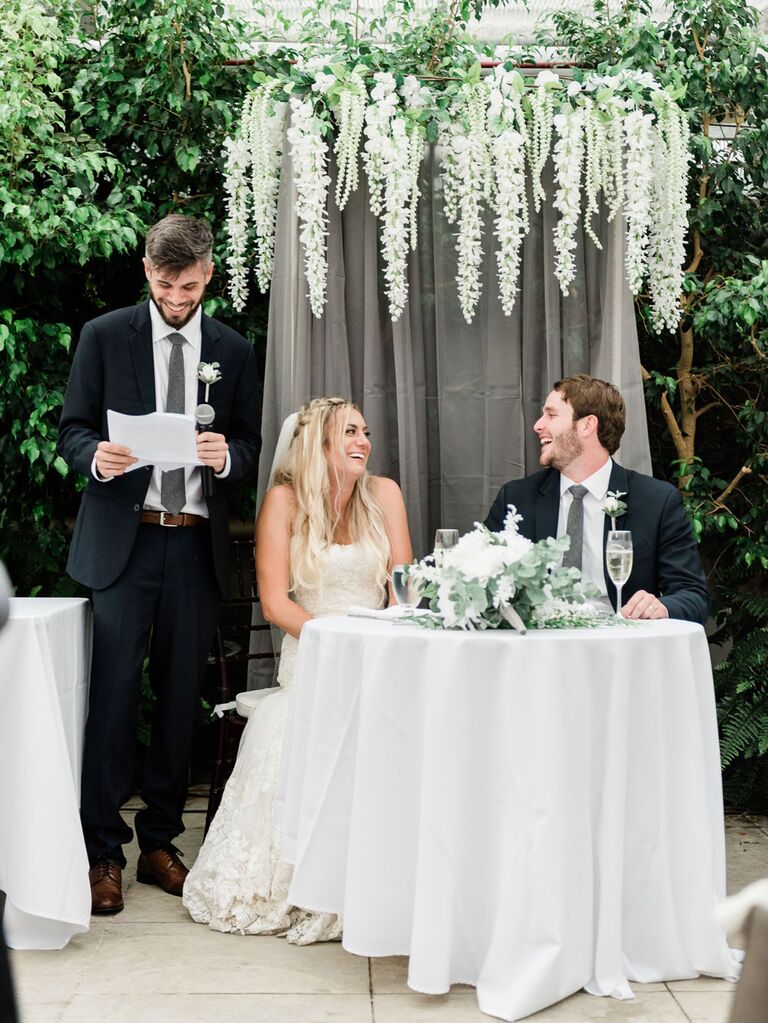 Best man giving toast at wedding reception while bride and groom laugh