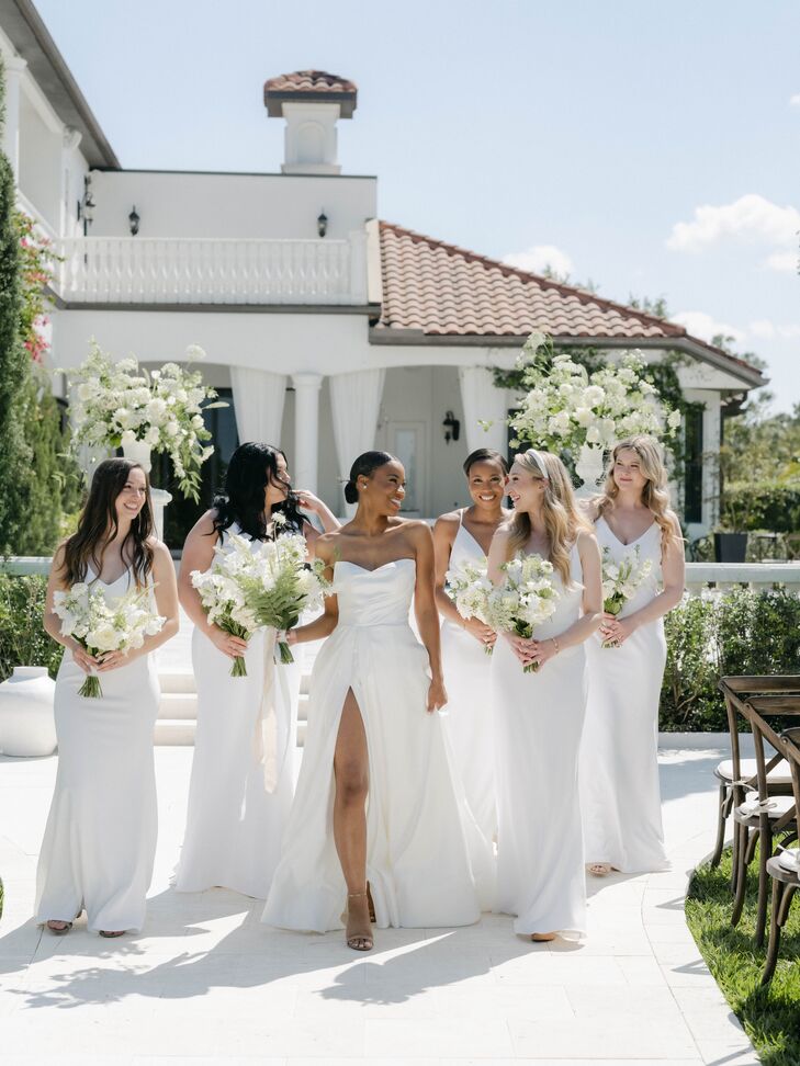 A bride in a sweetheart, A-line gown with a slit in the skirt and a low-bun hairstyle carries a bouquet of white flowers accents with greenery and poses with her bridesmaids in all-white dresses with their own white-and-green bouquets in an estate garden before her Italian garden-inspired wedding.